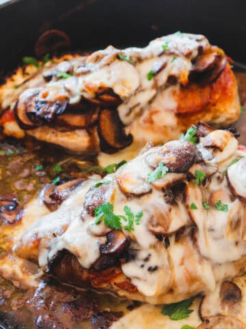 two chicken breasts smothered in mushrooms, onions, and melted cheese in a cast iron skillet.