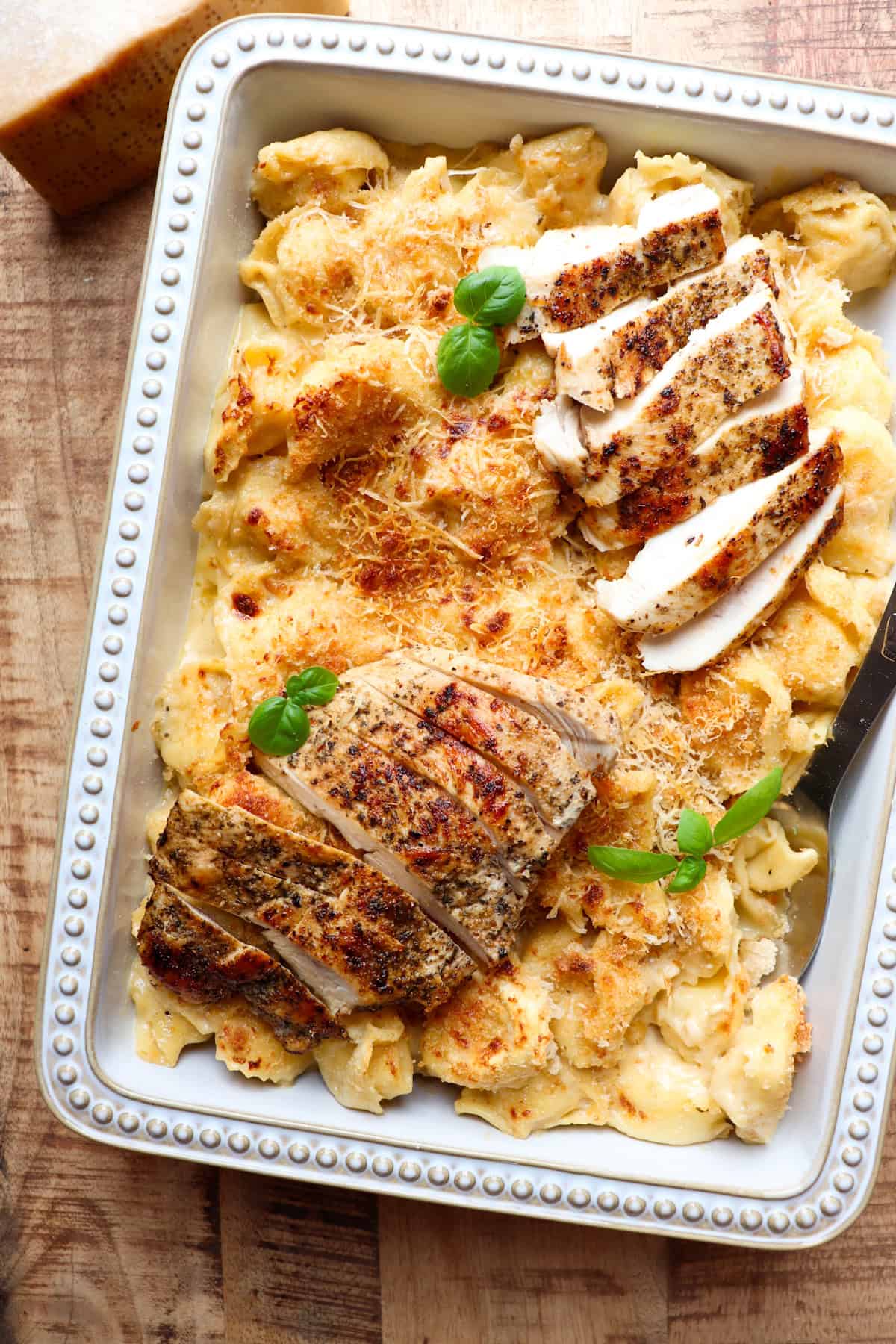 sliced chicken on top of a casserole dish of tortellini that have been baked in the oven with basil on top for garnish.