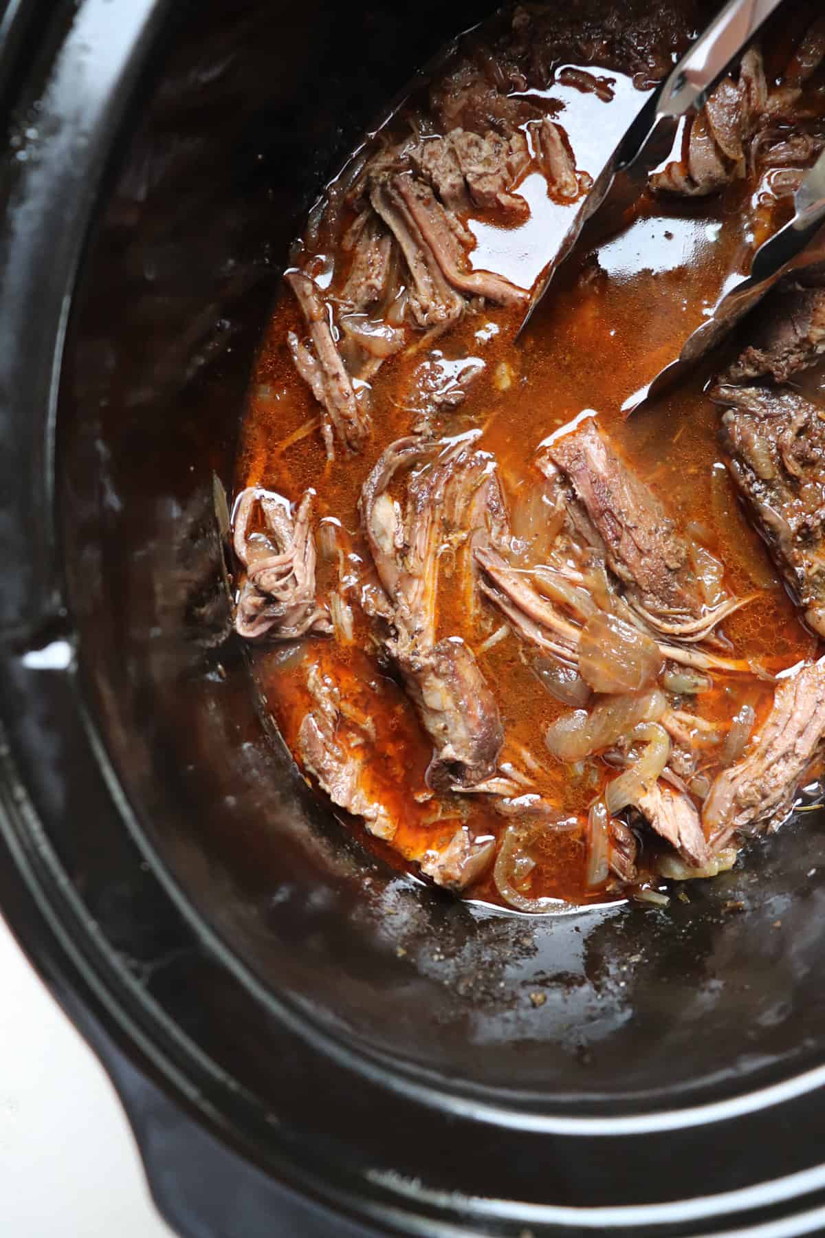 tongs grabbing meat from a black slow cooker.