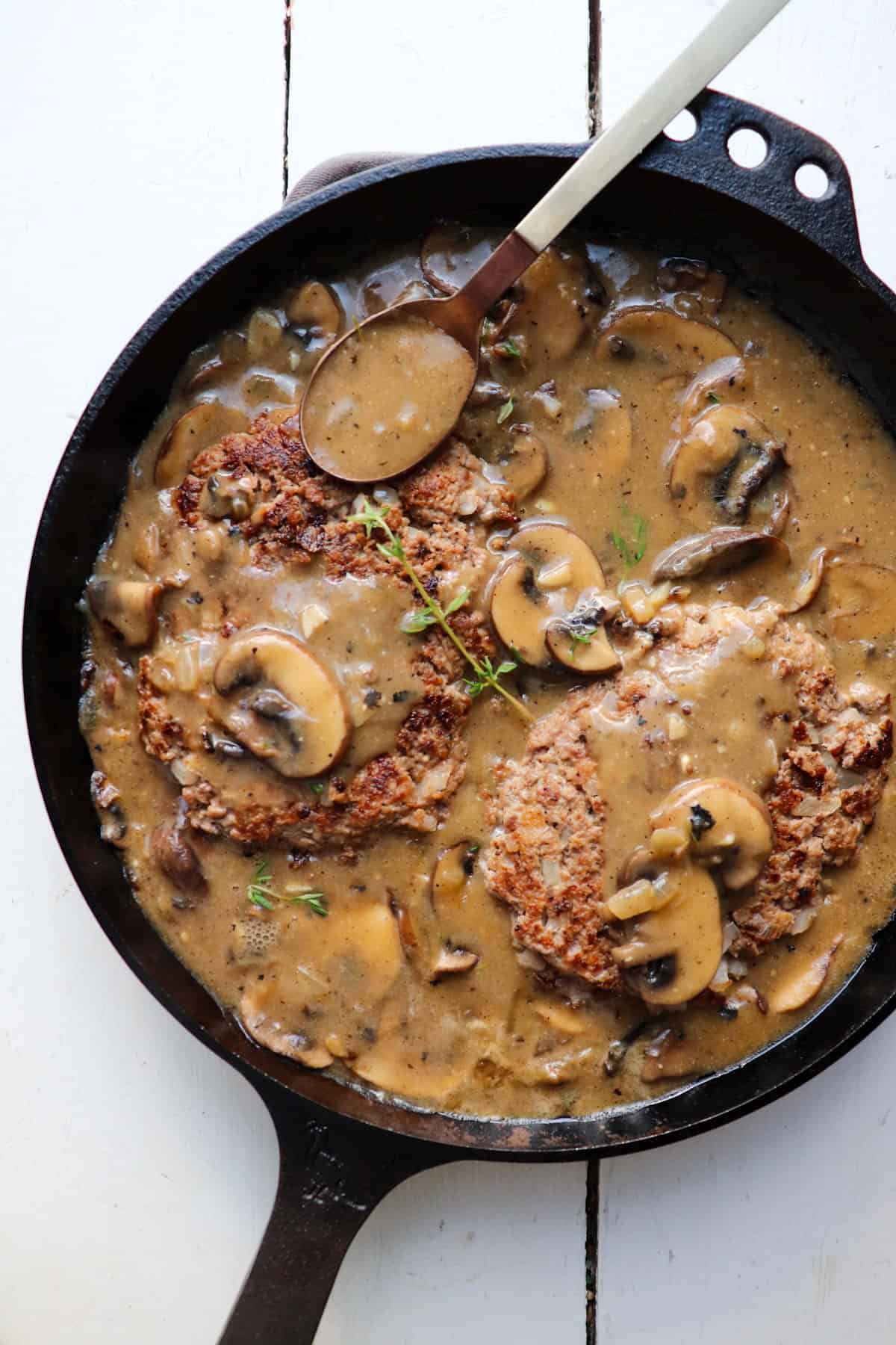 A skillet filled with salisbury steak patties and mushrooms with a spoon scooping the sauce.