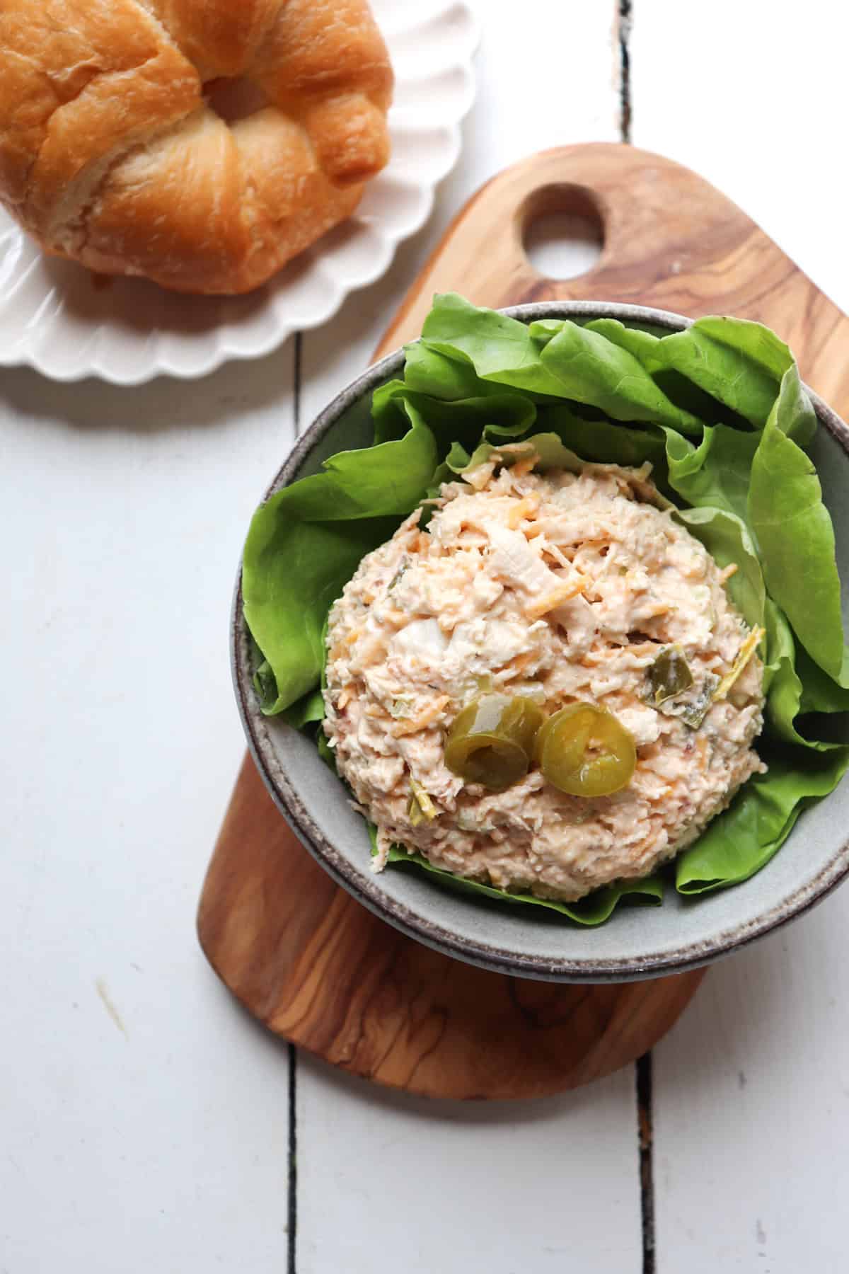 scoop of chicken salad with pickled jalapenos on top on a bed of lettuce with a croissant in the background.