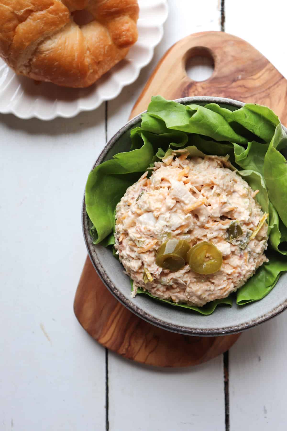 scoop of chicken salad with pickled jalapenos on top on a bed of lettuce with a croissant in the background.
