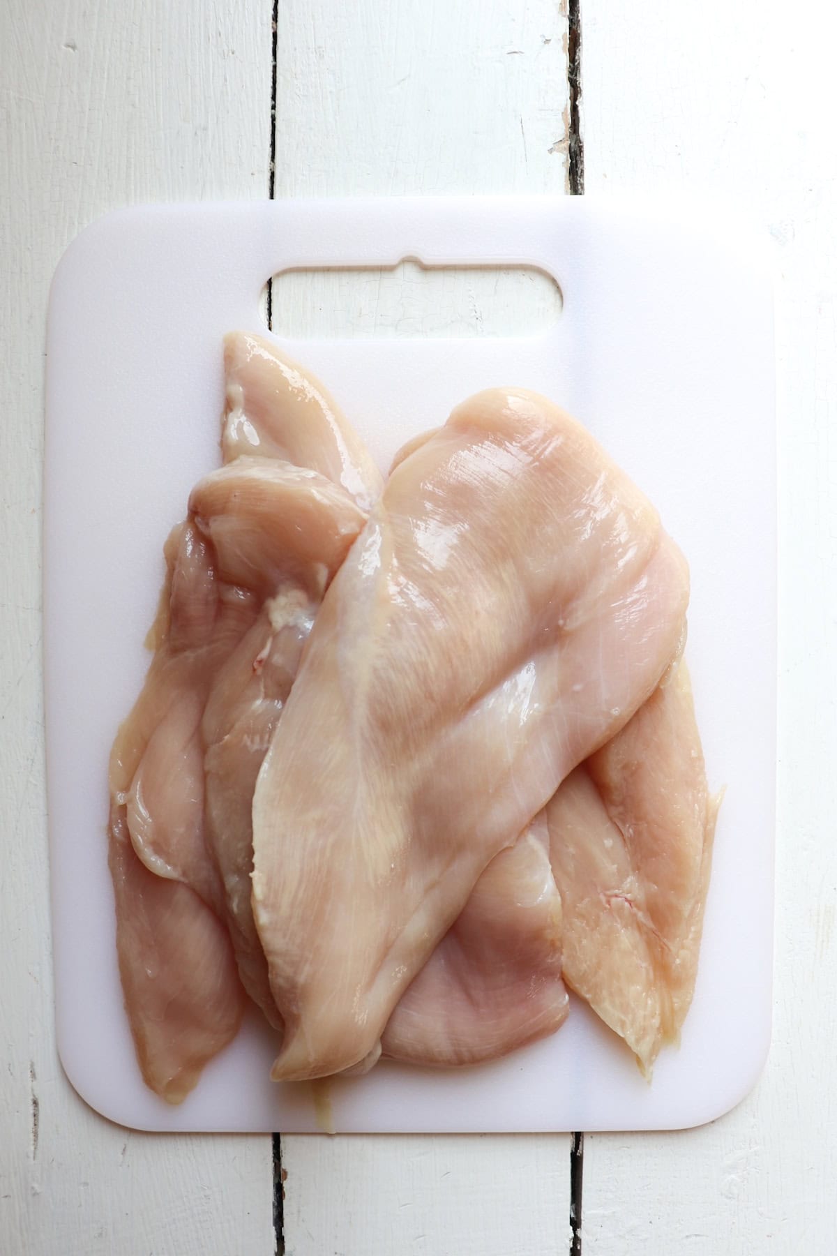 four raw chicken breast pieces on a white cutting board.