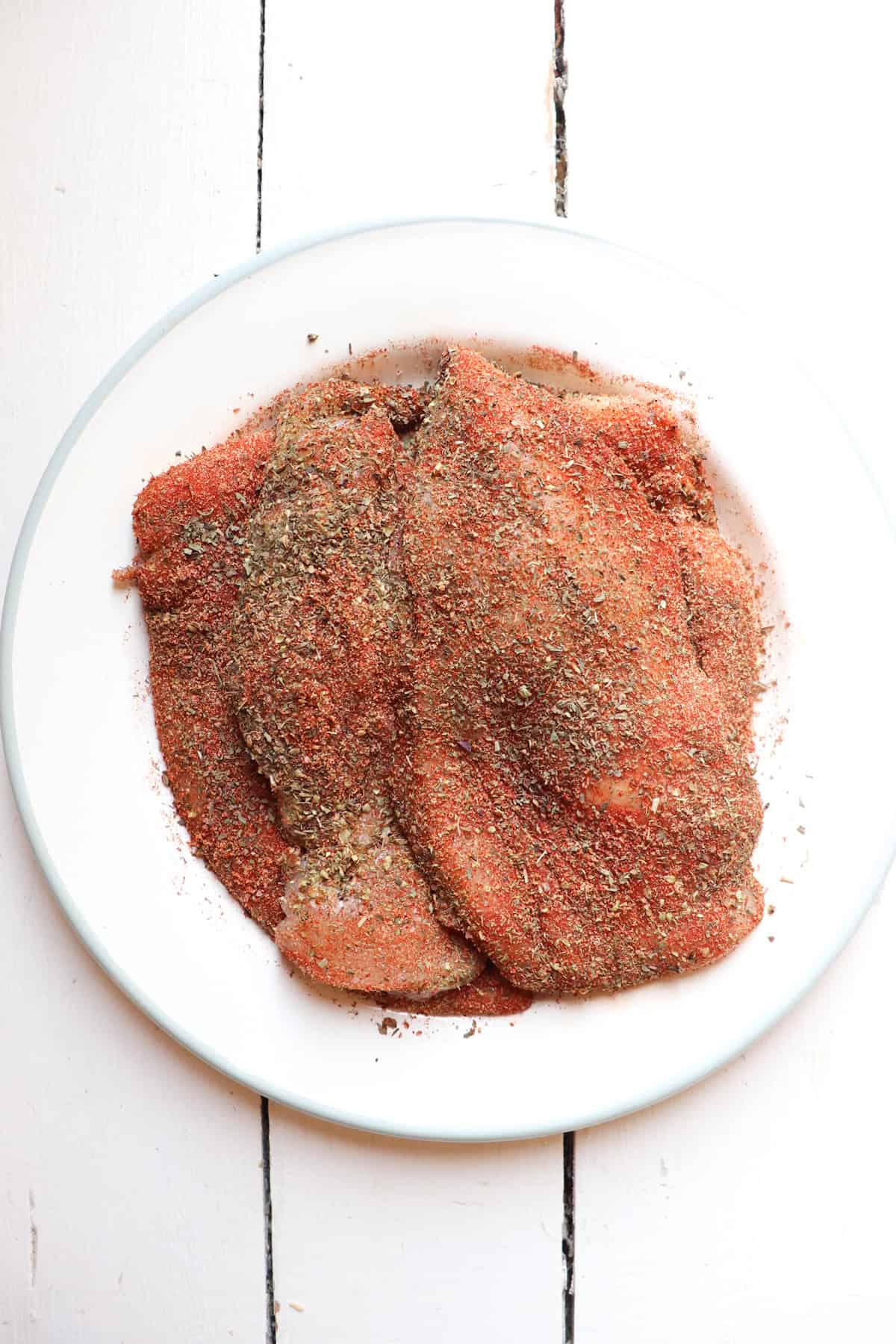 four thin cut chicken breasts coated in seasoning on a white plate.