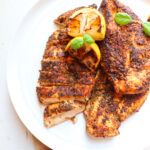 herb crusted chicken on a white plate with lemons and basil as garnish.