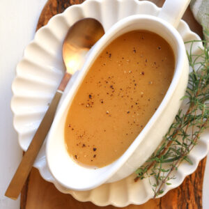 white gravy boat filled with gravy on a white plate with herbs and a spoon on the side.