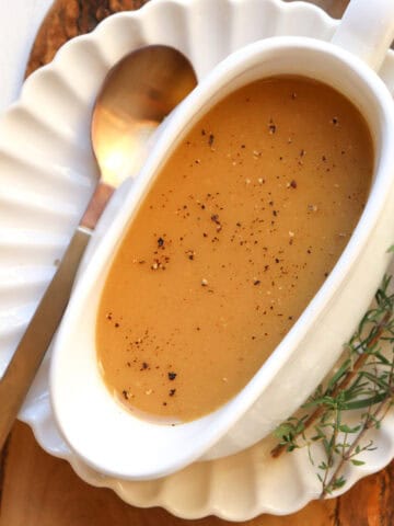 white gravy boat filled with gravy on a white plate with herbs and a spoon on the side.