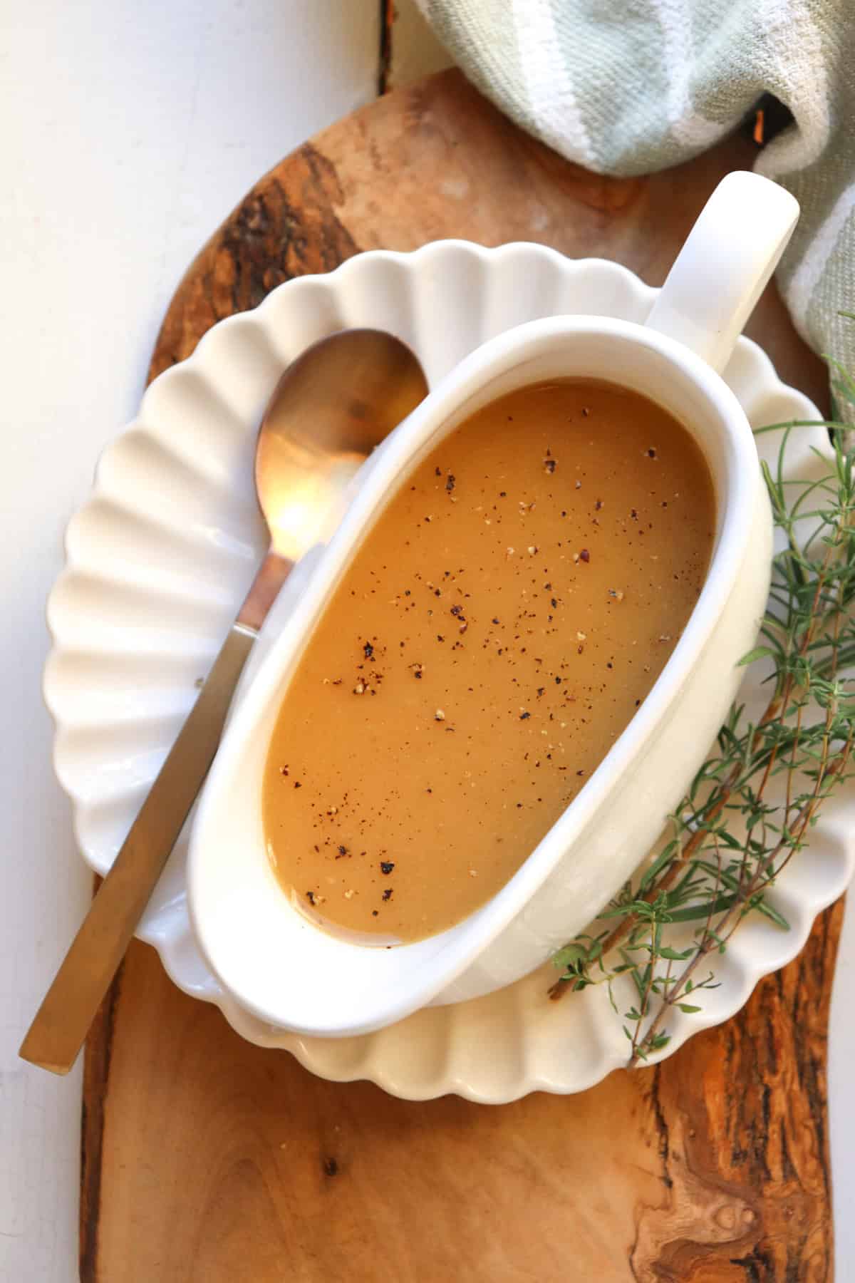 white gravy boat filled with gravy on a white plate with herbs and a spoon on the side.