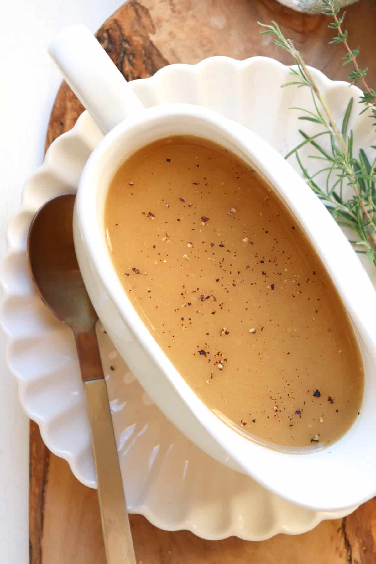 white gravy boat filled with gravy on a white plate with herbs on the side.