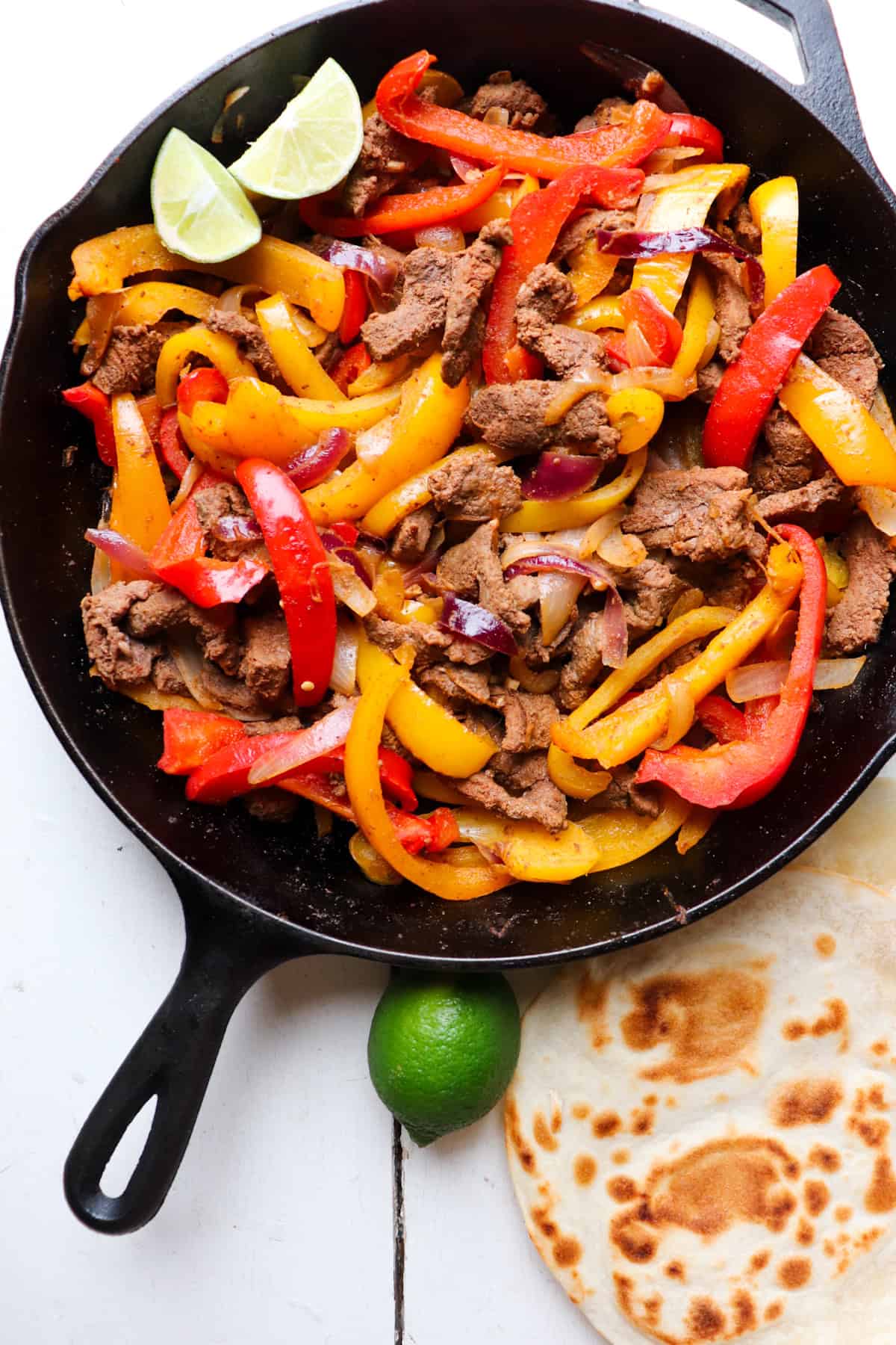 cast iron skillet filled with venison fajitas with tortillas and lime on the side.