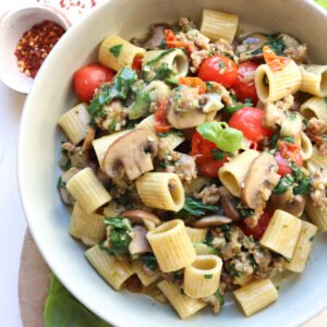 bowl of sausage and mushrooms pasta with red pepper flakes next to it.