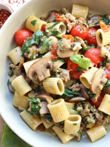 bowl of sausage and mushrooms pasta with red pepper flakes next to it.
