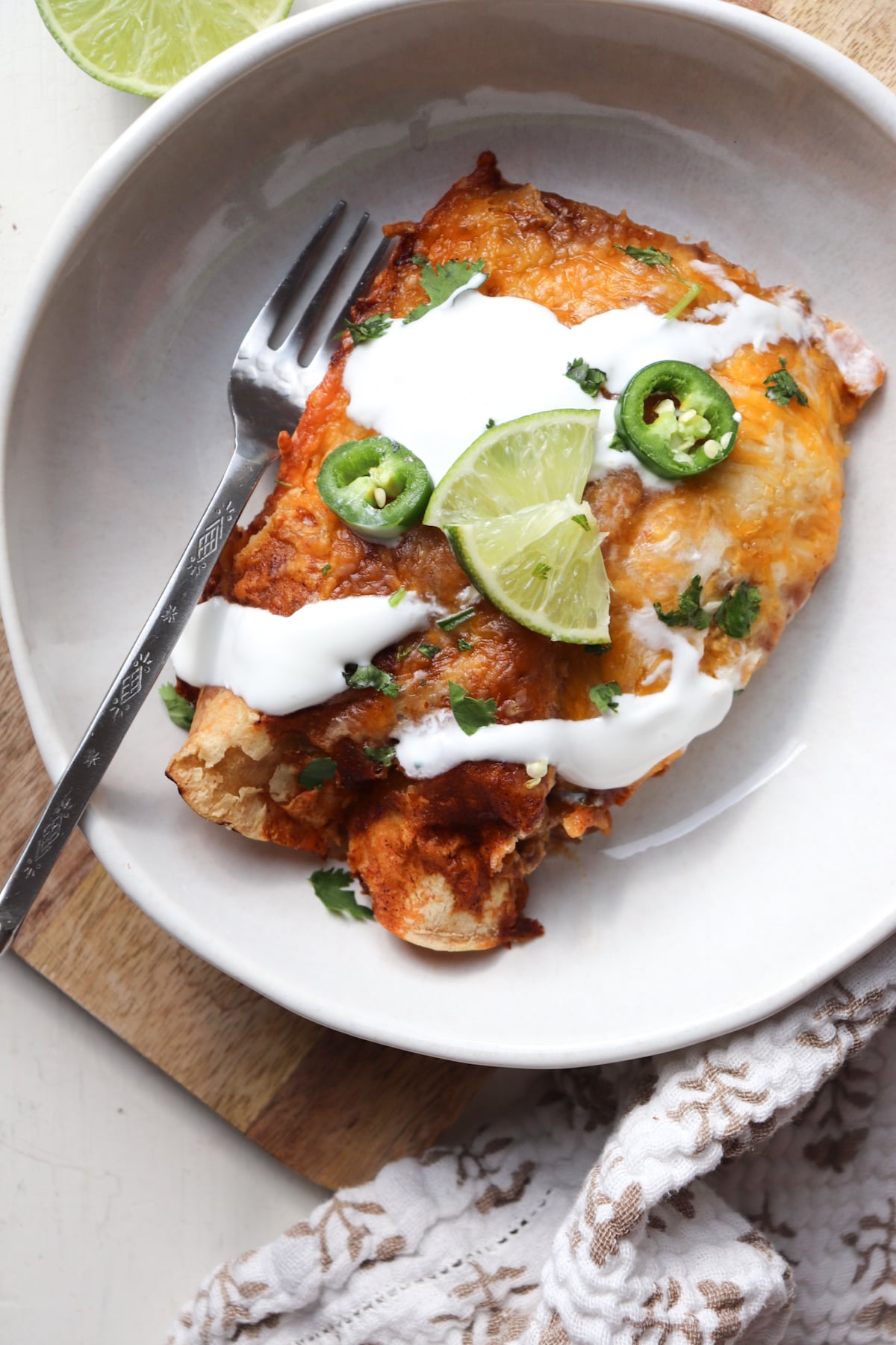plate with two enchiladas and topped with sour cream, limes, and jalapenos.